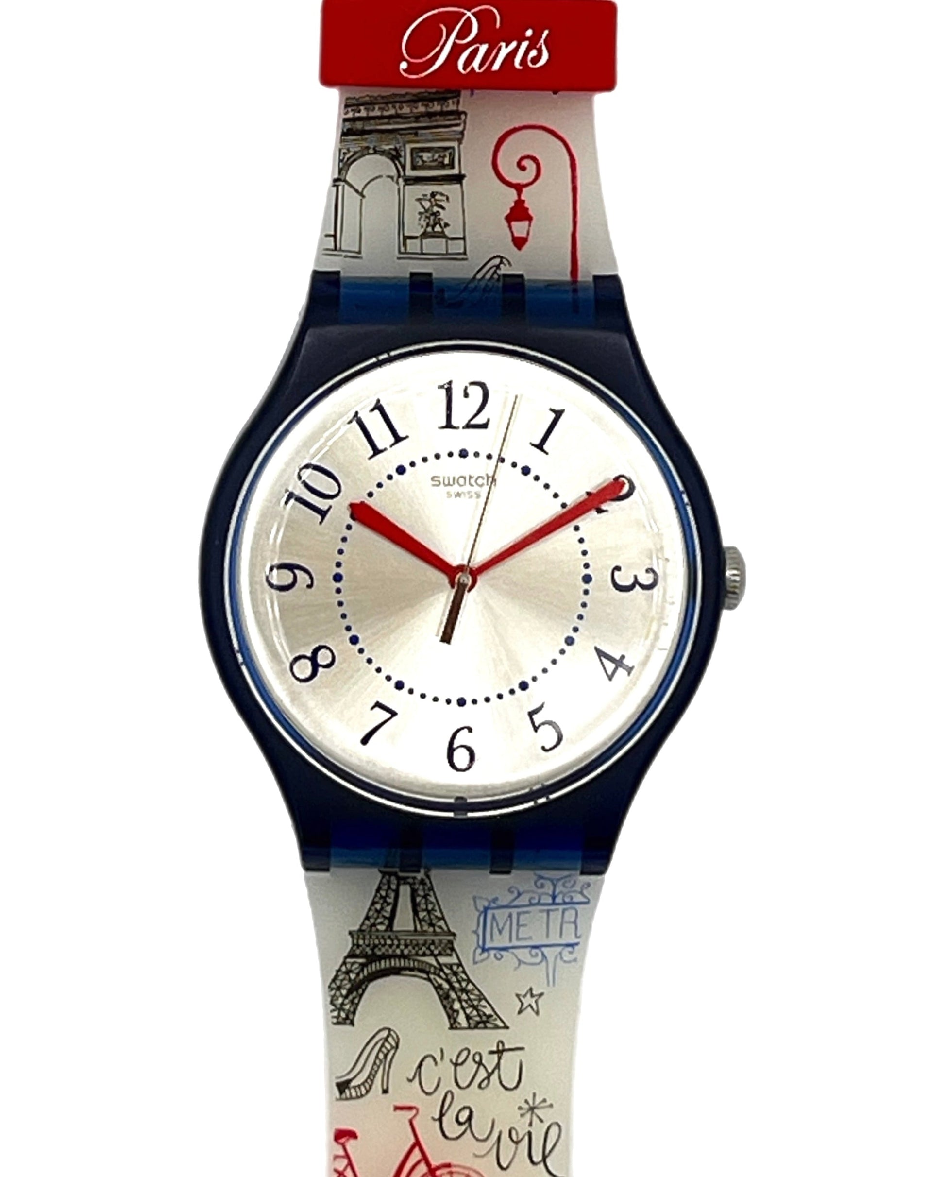 A Swatch Paris wristwatch featuring a white dial with red hands and black numerals, housed in a dark blue bezel. The strap is decorated with illustrations of the Eiffel Tower, a red bicycle, and 'Greetings from Paris' text, set against a plain white background.