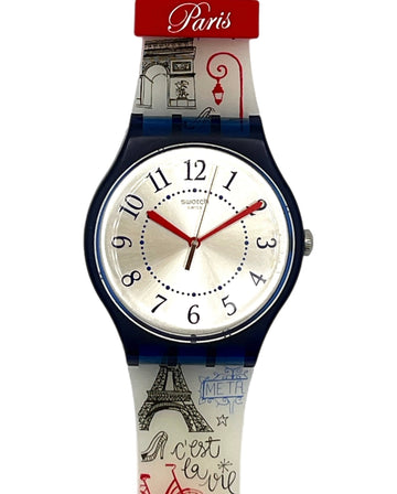 A Swatch Paris wristwatch featuring a white dial with red hands and black numerals, housed in a dark blue bezel. The strap is decorated with illustrations of the Eiffel Tower, a red bicycle, and 'Greetings from Paris' text, set against a plain white background.