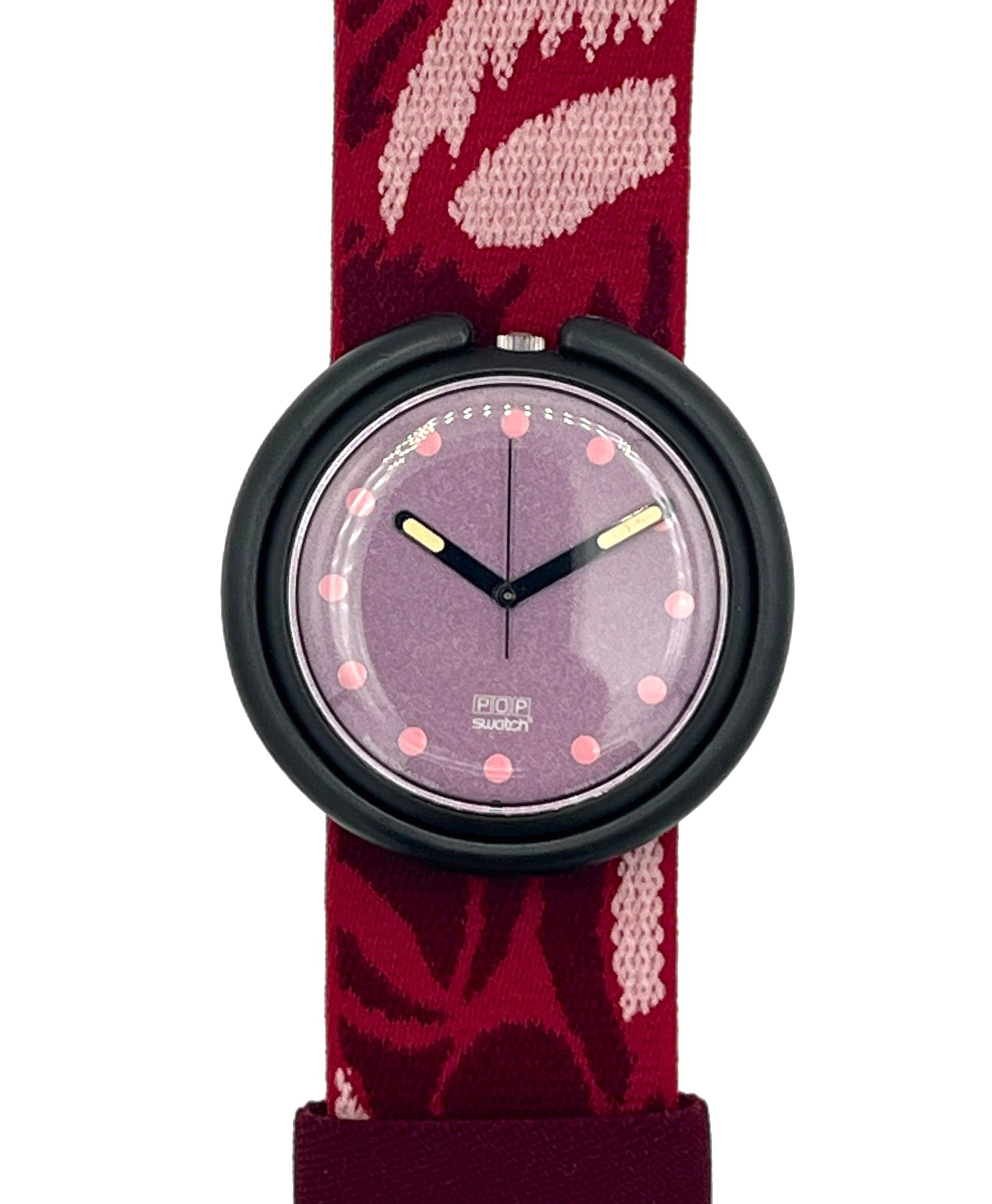 A Swatch wristwatch featuring a burgundy and pink patterned fabric strap, a black round bezel, and a lavender-purple dial with pink dot hour markers.