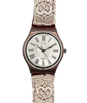 A vintage-style wristwatch with a round rose gold-toned case, a white dial with black Roman numerals, and a decorative off-white lace strap, shown against a plain white background.