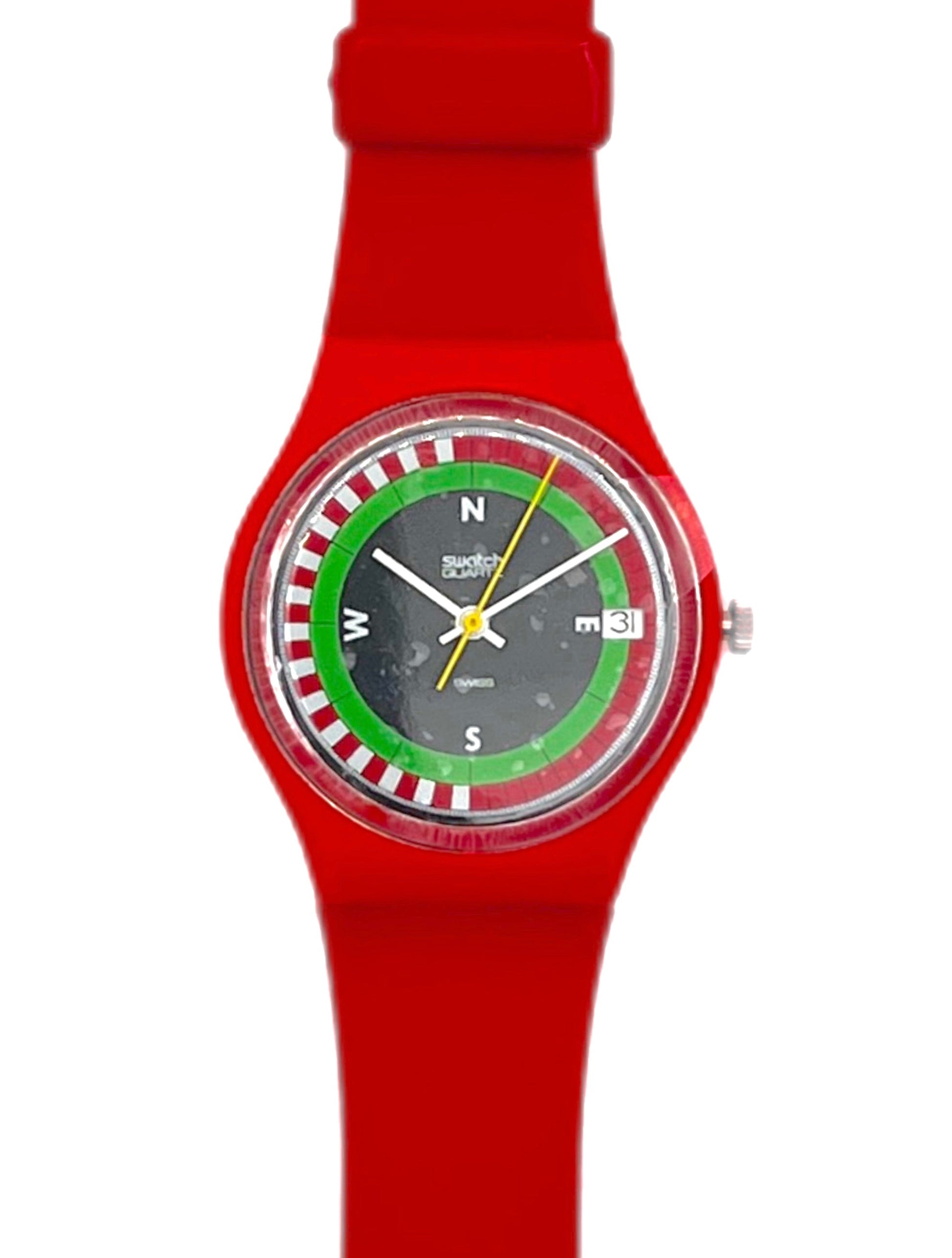 A Swatch wristwatch with a bright red silicone strap and case, featuring a unique dial with a green and red roulette wheel design, white letter markers, and yellow hands on a white background.