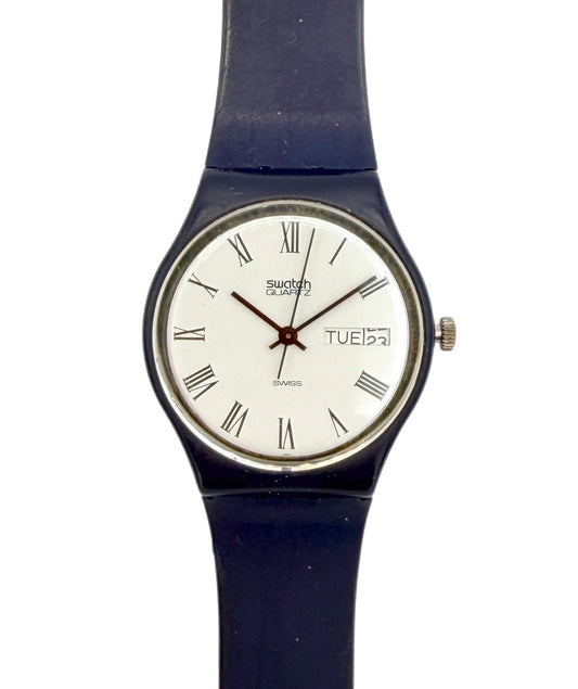 A Swatch wristwatch with a dark blue strap, a silver-tone case, and a white dial featuring black Roman numerals and a day window.