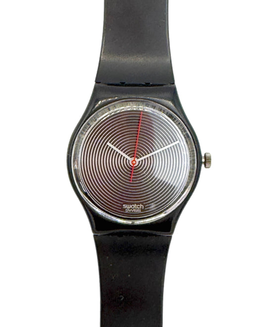 A Swatch wristwatch with a black strap, a silver-toned bezel, and a distinctive black-and-white concentric spiral dial with a red second hand.