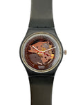 Swatch wristwatch with a black strap and case, featuring a transparent dial that reveals a copper-toned mechanical movement inside, shown against a plain white background.