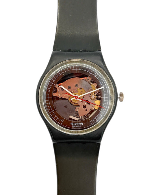 Swatch wristwatch with a black strap and case, featuring a transparent dial that reveals a copper-toned mechanical movement inside, shown against a plain white background.